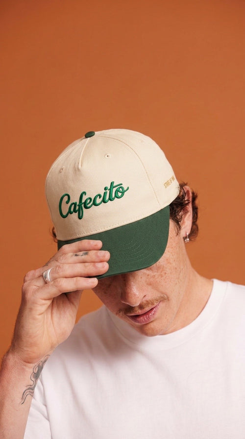 Person wearing a beige and green cap with 'Cafecito' text against an orange background